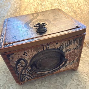 May include: A rectangular wooden box featuring a decorative surface with musical notes, a treble clef, and floral patterns. The lid has a small metal ring handle, while the front displays a large, oval-shaped antique bronze drop handle.