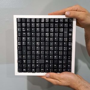 Pode incluir: Uma obra de arte emoldurada feita de teclas de teclado pretas, soletrando uma citação de John Lennon: "Conte sua idade por amigos, não por anos. Conte sua vida por sorrisos, não por lágrimas." A moldura é branca.
