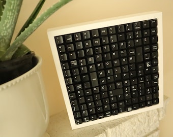 May include: A framed artwork made from black keyboard keys. The keys spell out the phrase "IF YOU ARE GOING THROUGH HELL KEEP GOING". The frame is white, and the artwork is displayed on a white brick surface.