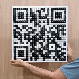 May include: A square artwork composed of black and white keyboard keys arranged in a QR code pattern, set within a white frame. The piece is held up against a wooden background.