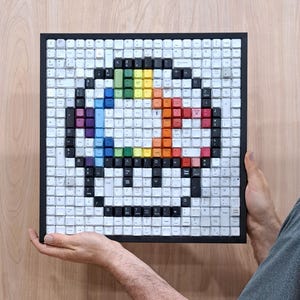May include: A framed artwork composed of white and colored keyboard keys. The keys are arranged to form a pixelated rainbow-colored design. The artwork is held by hands against a light wood background.