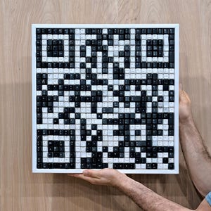 Pode incluir: Uma obra de arte quadrada composta por teclas de teclado pretas e brancas, dispostas para formar um código QR. As teclas estão dentro de uma moldura branca. A obra é segurada por duas mãos contra um fundo de madeira.