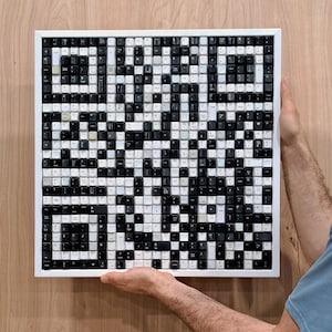 Pode incluir: Uma obra de arte emoldurada composta por teclas de teclado pretas e brancas, formando um design de código QR. As teclas são dispostas em um padrão de grade dentro de uma moldura branca. A obra de arte é segurada por uma pessoa.