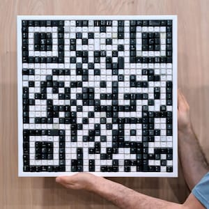 Pode incluir: Uma obra de arte quadrada composta por teclas de teclado pretas e brancas, soletrando a palavra "GUESS" em um estilo pixelizado. As teclas estão em uma moldura branca, criando um contraste marcante. A obra de arte é segurada por uma pessoa.