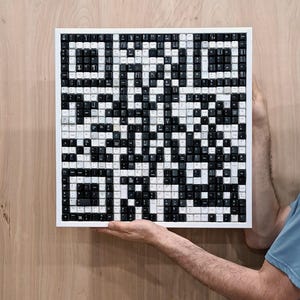 Pode incluir: Uma obra de arte quadrada com um design de código QR, construída a partir de teclas de teclado pretas e brancas. As teclas são dispostas dentro de uma moldura branca, criando um padrão de alto contraste. A obra é exibida sobre um fundo de madeira.