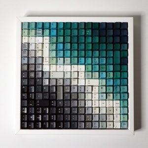 May include: A framed artwork composed of repurposed keyboard keys. The keys are arranged in a gradient of blues, greens, and grays, creating an abstract design. The white frame provides a clean border for the piece.