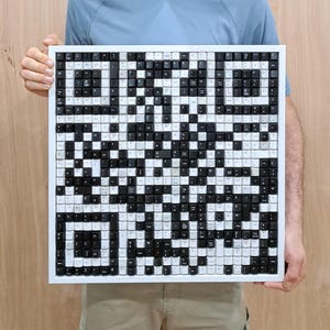 Pode incluir: Uma obra de arte quadrada feita de teclas de teclado recicladas. As teclas, pretas e brancas, são dispostas para criar um design de código QR. A obra de arte é emoldurada em uma borda branca.