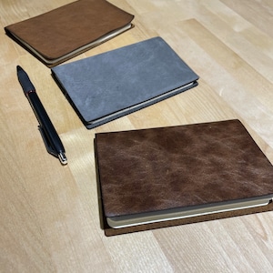 May include: Three leather-bound notebooks in brown, grey, and dark brown, lying on a wooden surface. A black pen is lying next to the notebooks.