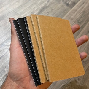 May include: A stack of four notebooks, two with black covers and two with brown covers. The notebooks are bound with stitching along the spine.