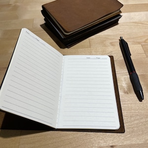 May include: A brown leather-bound journal with lined pages is open to a blank page. The journal is lying on a wooden surface with a black pen next to it.