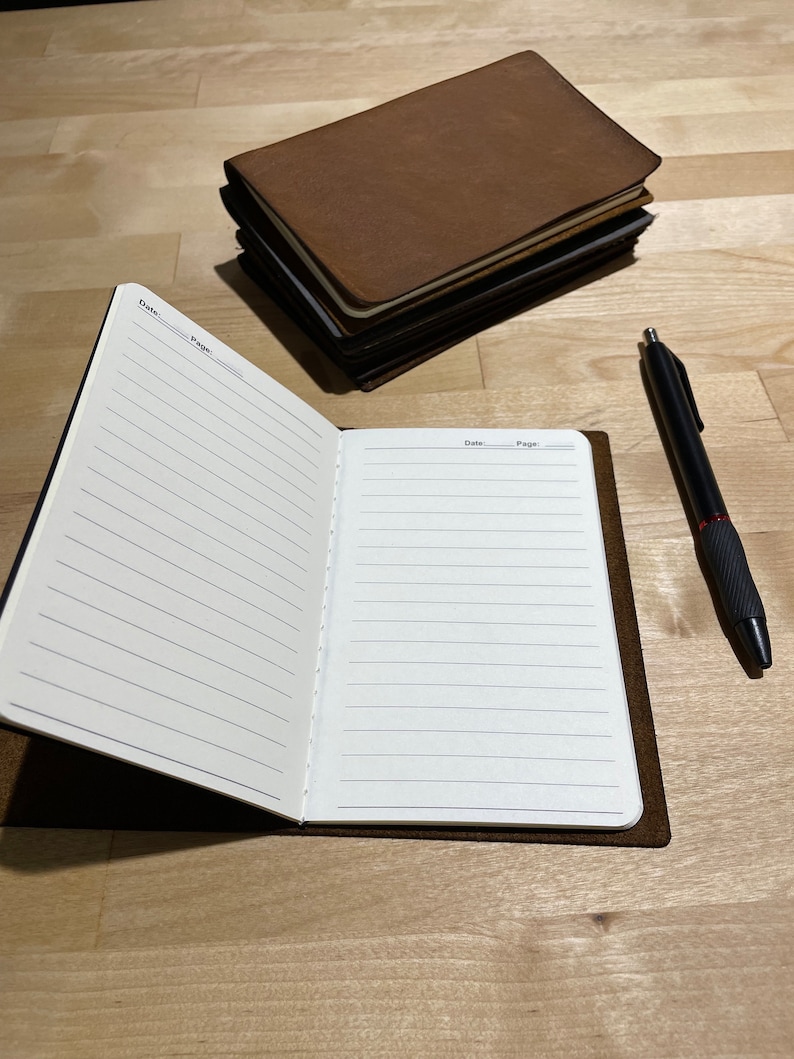 May include: A brown leather-bound journal with lined pages is open to a blank page. The journal is lying on a wooden surface with a black pen next to it.