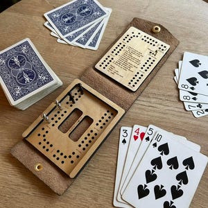 Puede incluir: Un tablero de cribbage de madera con una funda de cuero, naipes y una guía de puntuación de madera. Las cartas tienen dorsos azules y el tablero tiene agujeros para clavijas. La guía de puntuación tiene texto.