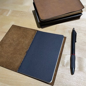 May include: A brown leather notebook with a black paper insert and a black pen. The notebook is open and the pen is lying on the table next to it.
