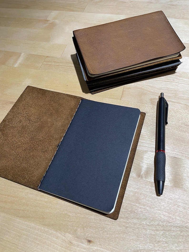 May include: A brown leather notebook with a black paper insert and a black pen. The notebook is open and the pen is lying on the table next to it.