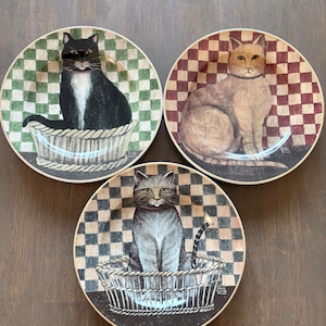 May include: Three decorative plates featuring cat illustrations. Each plate showcases a different cat breed sitting in a basket. The plates have checkered backgrounds in green, red, and black, with a brown rim.