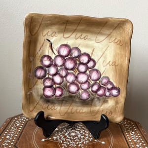 May include: Square decorative plate with a hand-painted bunch of purple grapes. The plate has a light brown background with the cursive text "Via" repeated across the surface. The plate is displayed on a black stand.