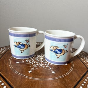 May include: Two white ceramic mugs with a blue and lavender band around the top and a cartoon rabbit illustration. The rabbit is wearing a blue jacket and flying with a bird. The mugs have a curved handle.