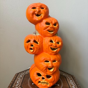 May include: A vertical stack of six orange ceramic pumpkins, each featuring a unique carved facial expression with cut-out eyes and mouths. The pumpkins are arranged in a column, with each pumpkin showing distinct, textured features and expressions.