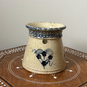 May include: A tan ceramic oil warmer with a blue heart and floral design. The rim is decorated with a blue speckled pattern. The warmer has a cylindrical shape with a flared base and several small holes.