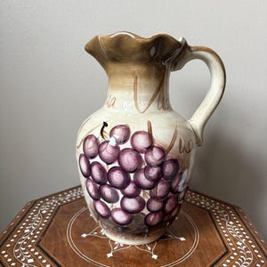 May include: A ceramic pitcher with a brown rim and handle, featuring a hand-painted design of purple grapes. The pitcher has a cream-colored body with the words "Via Vita" written on it. The pitcher is sitting on a wooden table.