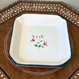 May include: A square white ceramic dish with rounded corners and a dark green rim. The center of the dish features a small, delicate illustration of green holly leaves and bright red berries on thin, light green stems against the white background.