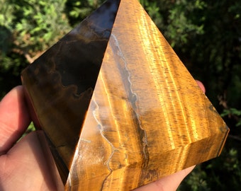 May include: A large, golden brown tiger's eye pyramid crystal with a smooth, polished surface. The crystal has a distinctive chatoyancy, creating a shimmering effect.