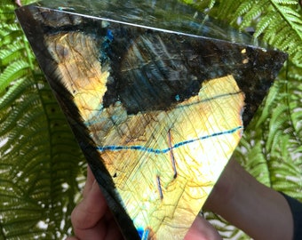 May include: A large, iridescent, pyramid-shaped labradorite crystal with a blue and gold color scheme. The crystal is smooth and polished, with a reflective surface.