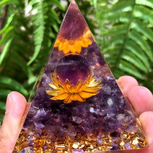 May include: A clear pyramid made of amethyst crystals with a gold flower and a purple amethyst sphere inside. The pyramid is surrounded by gold flakes.
