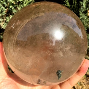 May include: A large, clear, smoky quartz crystal sphere. The sphere is smooth and polished, with a slight brown tint. It is being held in a hand.