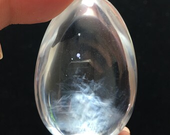 May include: A clear, egg-shaped crystal with internal white and blue feathery inclusions. The crystal is held up against a dark background, showcasing its transparency and the delicate patterns within. The crystal is smooth and polished.