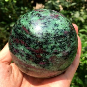 Puede incluir: Una esfera de piedra preciosa verde, negra y rosa. La esfera está pulida y tiene una superficie lisa.