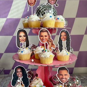 May include: A three-tiered pink cupcake stand with cupcakes decorated with colourful sprinkles. Each cupcake has a face cutout topper of a different person.