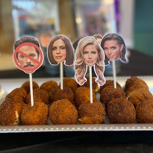 May include: A silver tray with a variety of small, round, brown, and crispy snacks. The snacks are topped with decorative paper cutouts of faces.