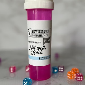May include: A pink prescription bottle with a white lid and a label that reads "BRAVOCON 2025 NOVEMBER 14-16". The label also says "Not well, Bitch" and "RECOVERY KIT". Scattered around the bottle are colorful dice.