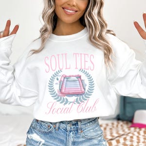 May include: White sweatshirt with a pink graphic design that says "Soul Ties Social Club" and features a pink and blue inflatable pool float.