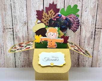May include: A handmade pop-up Thanksgiving card featuring a turkey, scarecrow, and autumn leaves. The card is primarily yellow, brown, green, and red. The front of the card has the words "Many Thanks" in elegant script.