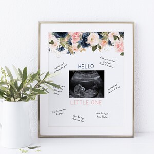 Puede incluir: Un marco blanco con un borde dorado sostiene una tarjeta blanca con un diseño floral en la parte superior. La tarjeta tiene una imagen de ultrasonido en blanco y negro en el centro con la palabra "HELLO" encima y las palabras "LITTLE ONE" debajo. Hay varios mensajes escritos a mano alrededor de la imagen de ultrasonido.