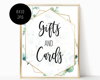 May include: A white sign with a gold geometric frame and green eucalyptus leaves. The sign says "Gifts and Cards" in black calligraphy.