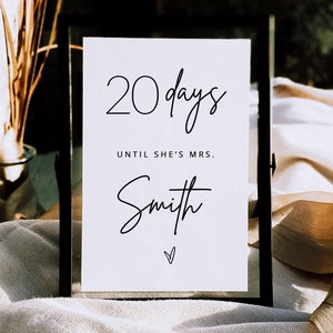 May include: A white sign with black text that reads "20 days until she's Mrs. Smith" with a small heart at the bottom. The sign is in a black frame and is sitting on a white surface.