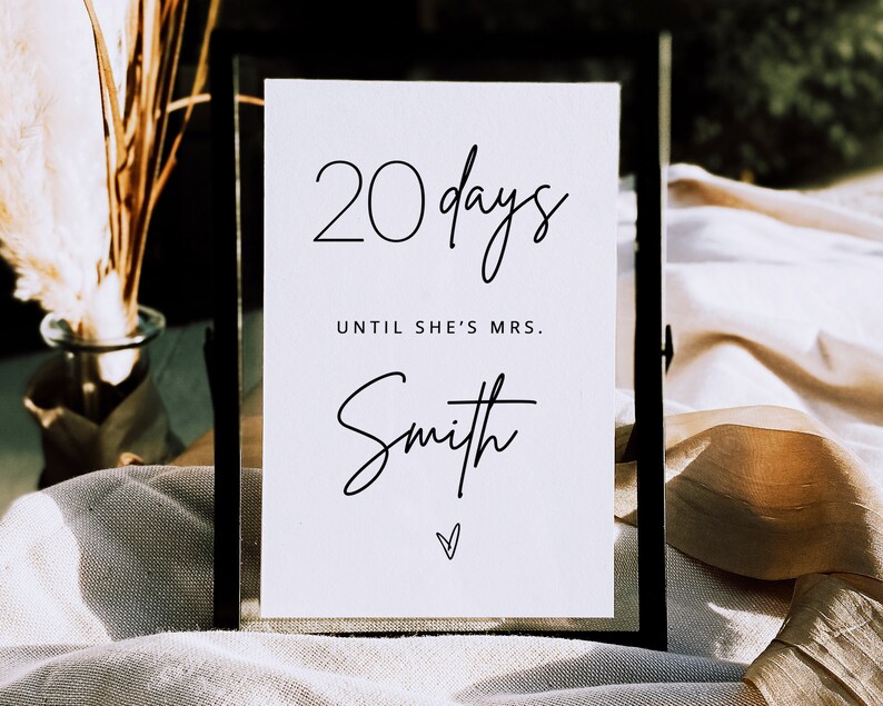May include: A white sign with black text that reads "20 days until she's Mrs. Smith" with a small heart at the bottom. The sign is in a black frame and is sitting on a white surface.