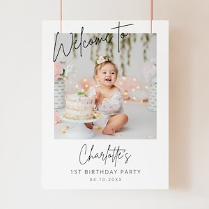 May include: Welcome to Charlotte's 1st Birthday Party, a white sign with a photo of a baby girl in a white dress sitting in front of a cake. The sign has black text and a white border.