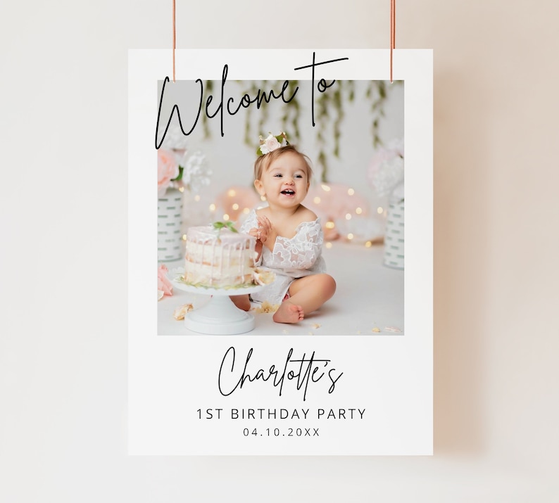 May include: Welcome to Charlotte's 1st Birthday Party, a white sign with a photo of a baby girl in a white dress sitting in front of a cake. The sign has black text and a white border.
