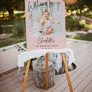 May include: Pink welcome sign with a photo of a baby girl in a white dress holding a cake. The sign says "Welcome to Charlotte's 1st Birthday Party 04.10.20XX".