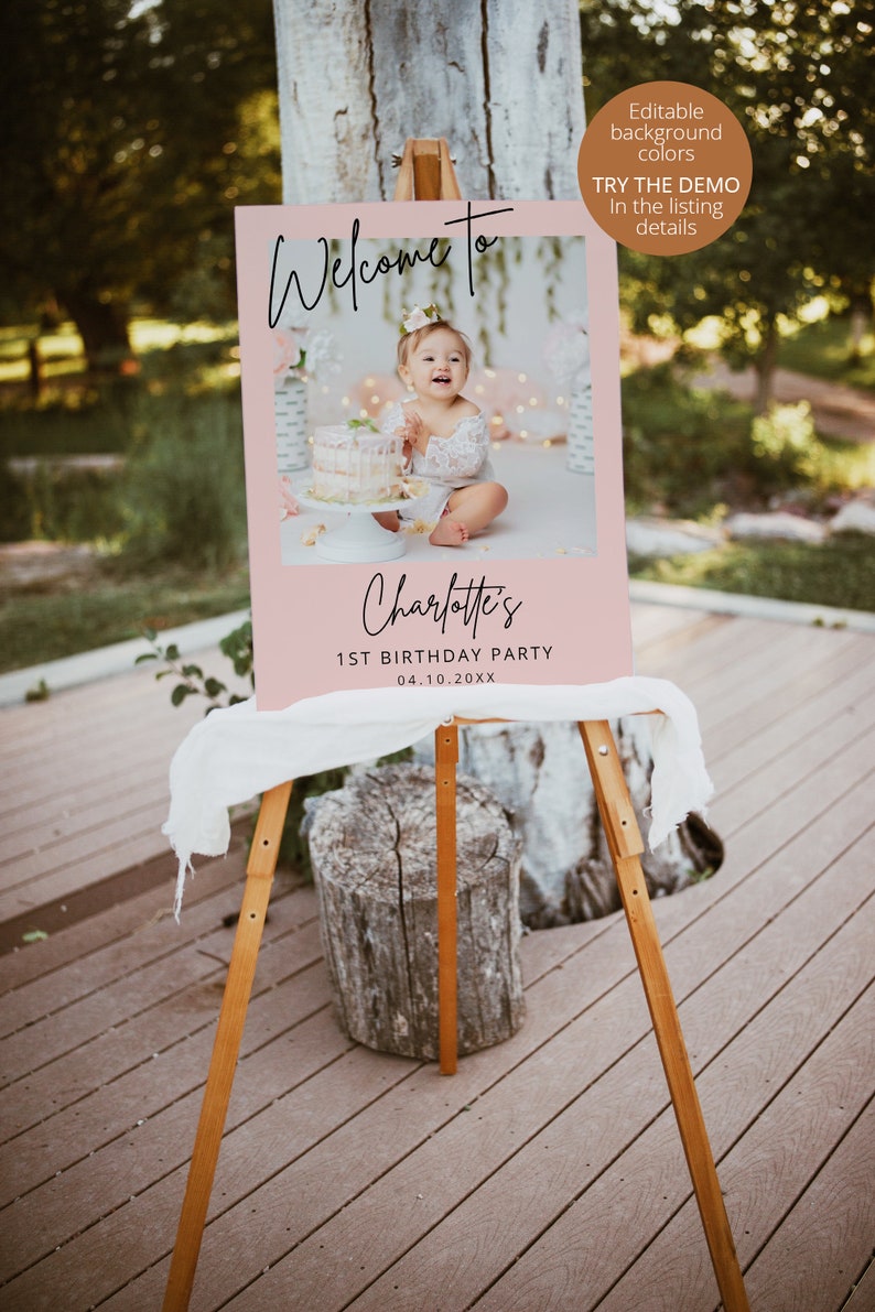 May include: Pink welcome sign with a photo of a baby girl in a white dress holding a cake. The sign says "Welcome to Charlotte's 1st Birthday Party 04.10.20XX".