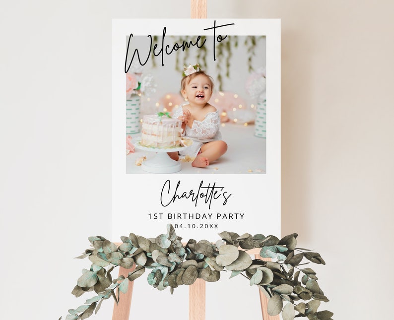 May include: White welcome sign with a photo of a baby girl in a white dress holding a cake. The sign says "Welcome to Charlotte's 1st Birthday Party 04.10.20XX"