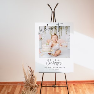 May include: Welcome to Charlotte's 1st Birthday Party sign with a photo of a baby girl in a white dress sitting in front of a cake. The sign is on an easel.