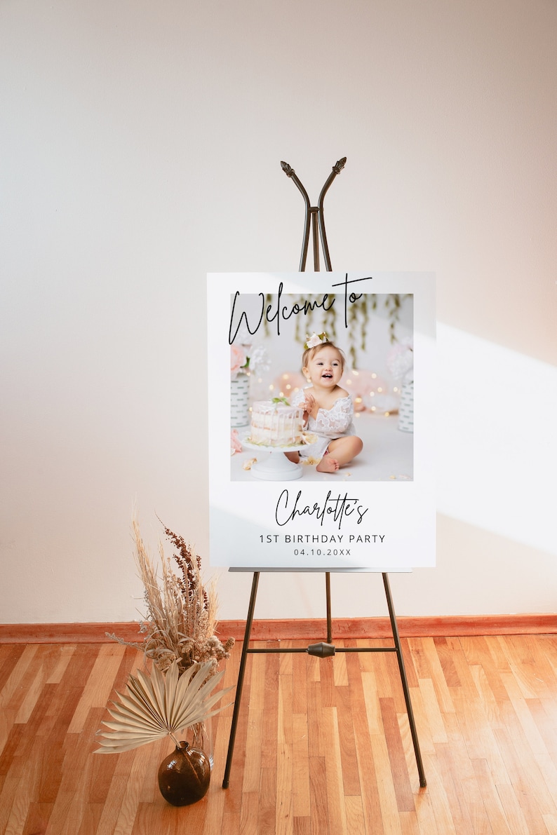 May include: Welcome to Charlotte's 1st Birthday Party sign with a photo of a baby girl in a white dress sitting in front of a cake. The sign is on an easel.