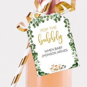 May include: A white tag with green foliage border and gold lettering that says "Pop the bubbly" and "When baby Jhonson arrives".  The tag is attached to a clear glass bottle with a gold and white striped straw.  There are small woodland animals at the bottom of the tag.