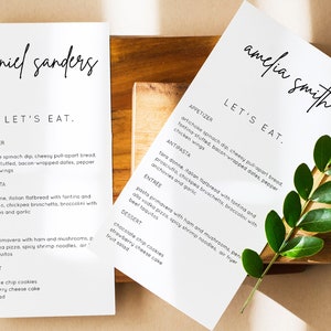 Puede incluir: Dos men&uacute;s blancos con texto negro, uno para Daniel Sanders y otro para Amelia Smith. Ambos men&uacute;s tienen los mismos platos enumerados: aperitivo, antipasto, plato principal y postre. Los men&uacute;s est&aacute;n sobre una superficie de madera con una planta verde.