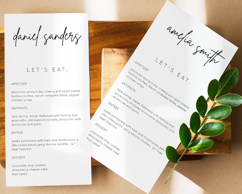 Puede incluir: Dos men&uacute;s blancos con texto negro, uno para Daniel Sanders y otro para Amelia Smith. Ambos men&uacute;s tienen los mismos platos enumerados: aperitivo, antipasto, plato principal y postre. Los men&uacute;s est&aacute;n sobre una superficie de madera con una planta verde.